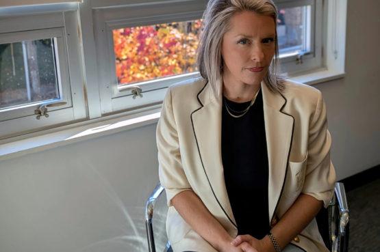 Woman in a white blazer seated by a window with colorful autumn foliage visible outside.